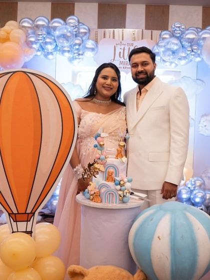 The couple stands beside their beautifully themed cake, surrounded by hot air balloon props and pastel balloon clouds. Every element is coordinated to bring the 'Up, Up & Away' theme to life.