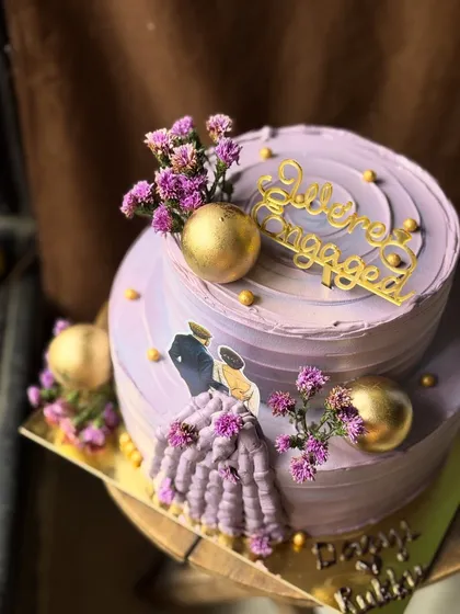 A stunning two-tier engagement cake in a lavender hue, adorned with fresh flowers and gold accents.