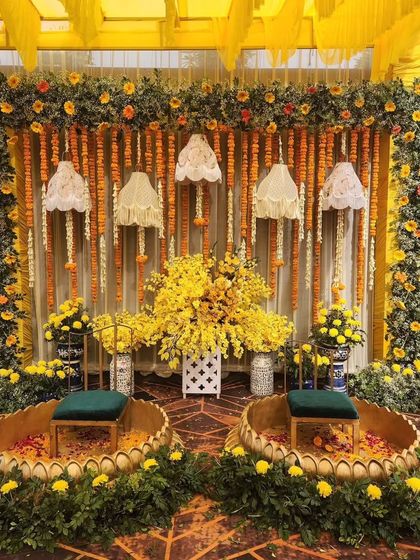 A traditional and vibrant haldi setup. The decor features two golden lotus-shaped seats, a backdrop of marigold and tuberose garlands, and hanging lace lampshades.