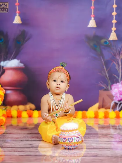 A classic pose for a Bal Krishna photoshoot, sitting with his flute and a pot of butter. The purple background adds a touch of royalty to the scene.