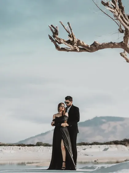 A dramatic and modern pre-wedding portrait set against a stark, beautiful landscape. The composition uses a bare tree to frame the couple, creating a powerful, high-fashion image that feels both artistic and romantic.