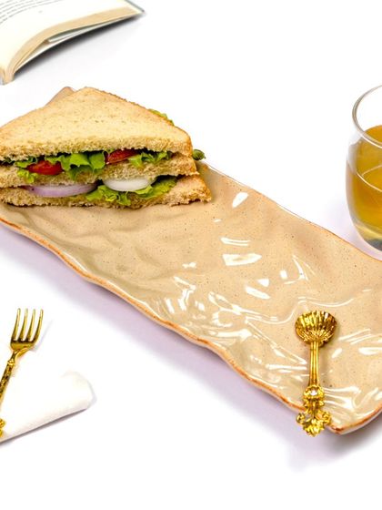 A long ceramic platter is styled for a simple lunch, holding a sandwich next to a glass of juice. The clean, bright lighting and minimal props create a fresh and appetizing scene.