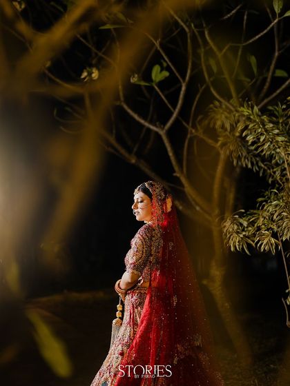 A stunning bridal portrait taken at night in Goa. The bride, in her crimson wedding lehenga, is illuminated against a backdrop of trees, creating a dramatic and enchanting image.
