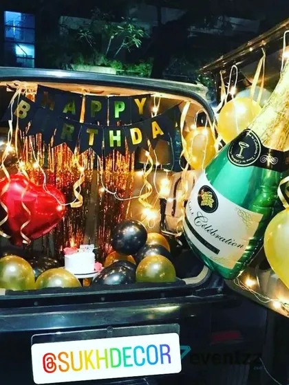 For a fun and unique surprise, I offer car boot decorations. This setup features a 'Happy Birthday' banner, a shimmering backdrop, festive gold and black balloons, and a large champagne bottle foil balloon to get the celebration started anywhere.
