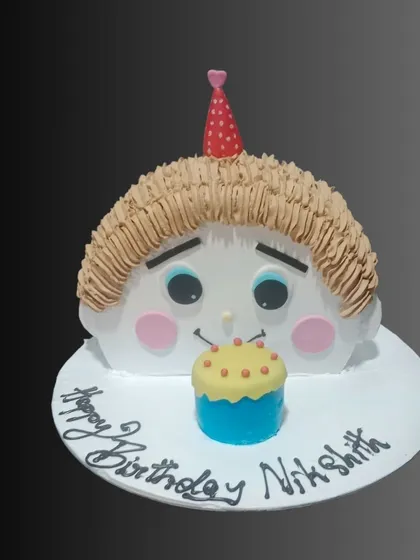 A cute and quirky custom cake sculpted to look like a cartoon baby's face, complete with a tiny birthday hat and a cupcake.