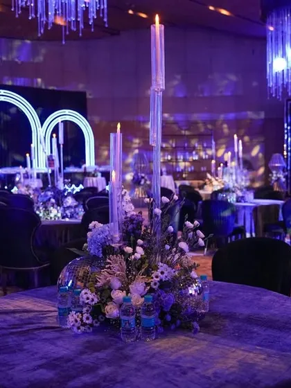A view of the dining area, with tables dressed in grey velvet and adorned with floral and candle centerpieces, looking towards the neon-arched cocktail bar.