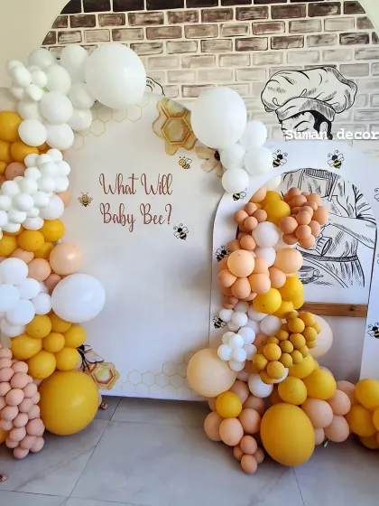 A fun "What will Baby Bee?" gender reveal party theme. The decoration includes a custom backdrop and a balloon arch in yellow, white, and peach colors.