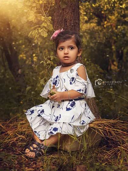A sweet portrait of a toddler girl sitting at the base of a tree in a sunlit forest. The natural setting provides a beautiful and peaceful backdrop.