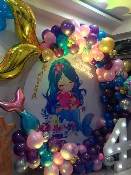 An underwater mermaid theme for a 4th birthday. The circular backdrop features a cute mermaid illustration, framed by a balloon garland with foil mermaid tail accents and a light-up number '4'.