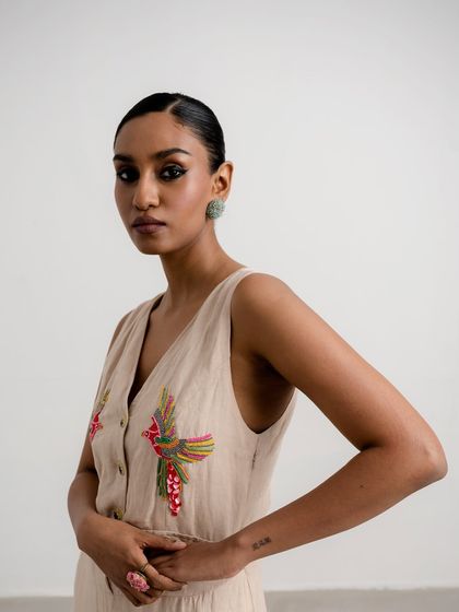 A close-up of a beige vest with colorful bird embroidery. The styling is minimal to highlight the playful and artistic details of the piece.