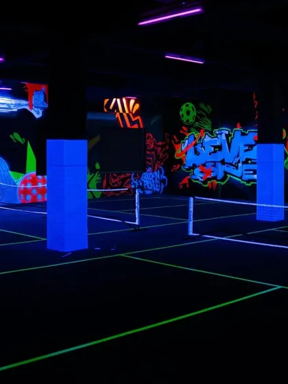 A wide-angle view of the entire pickleball court glowing under UV blacklights. The neon lines of the court and the glowing murals work together to create a completely immersive and futuristic playing environment.
