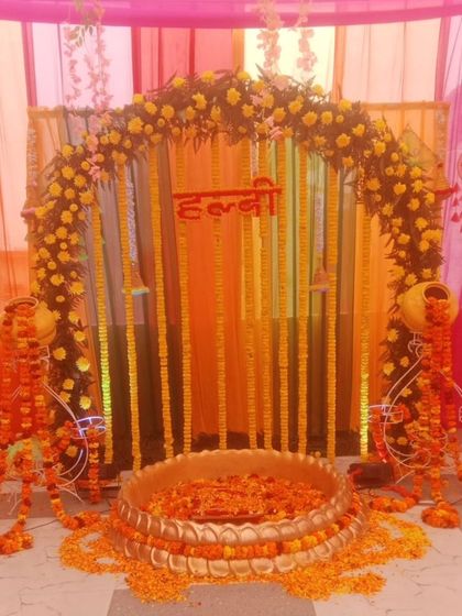 A traditional Haldi ceremony setup with a floral arch and a large brass urli. The decor uses marigold garlands and decorative matkas on stands, perfect for a home-based function.