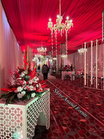 A striking red-themed entrance corridor with intricate floor patterns, floral arrangements, and elegant chandeliers hanging from the draped ceiling.