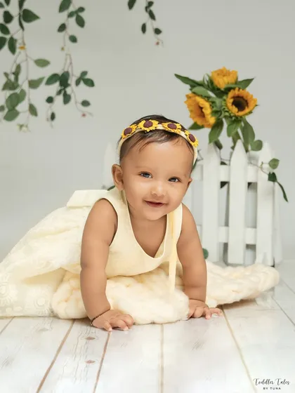This little girl, wearing a sunflower headband, gives a sweet, direct-to-camera smile. The simple white fence and greenery in the background create a lovely, fresh portrait.