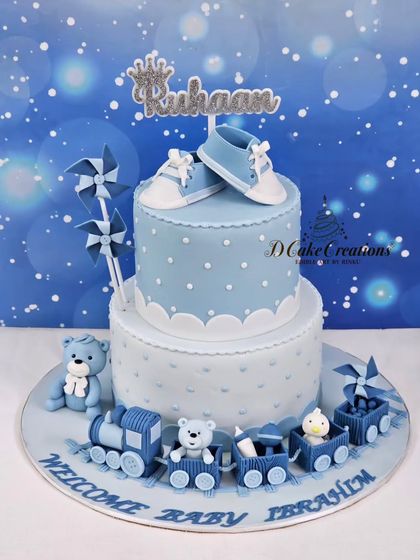 A studio shot of the "Welcome Baby" cake for Ruhaan. This two-tier creation features edible baby shoes, pinwheels, and a sweet teddy bear train.