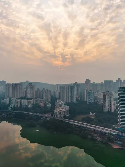 A stunning aerial view of the Powai skyline at sunrise. The clouds are lit up in gold, and their reflection is visible in the calm waters of the lake.
