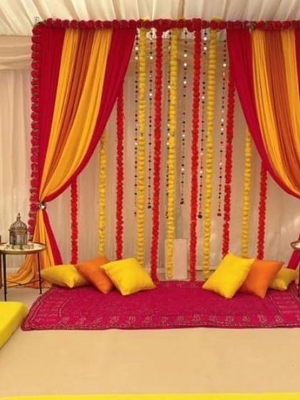 A comfortable and colorful low-seating arrangement for a Mehandi or Sangeet night. The backdrop of red and yellow drapes with marigold strings creates a warm and inviting atmosphere for guests.
