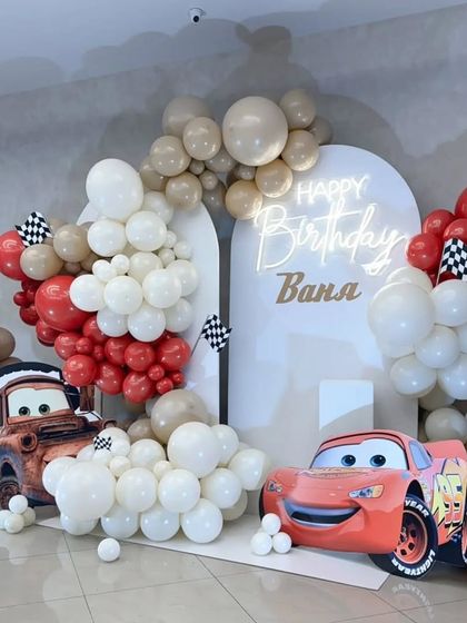 A modern take on the Disney Cars theme with a neutral color palette. The white arched backdrops are decorated with red, white, and beige balloons and character cutouts.