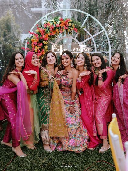 The bride, Arya, posing with her bridesmaids. The vibrant pinks and yellows of their outfits create a beautiful, coordinated look for the Mehendi or Sangeet.
