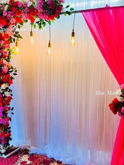 A colorful floral arch with hanging Edison bulbs, perfect for a birthday or anniversary party. The pink and red flowers create a vibrant and festive look.