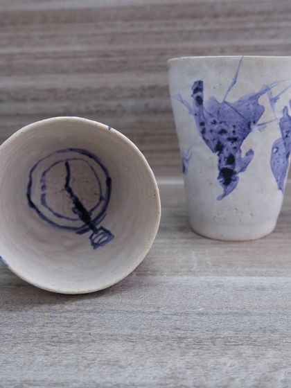 A set of two hand-painted ceramic glasses. These feature spontaneous, abstract blue designs on a white background, making each one a unique piece of functional art. They are microwave-safe for your milk or water.