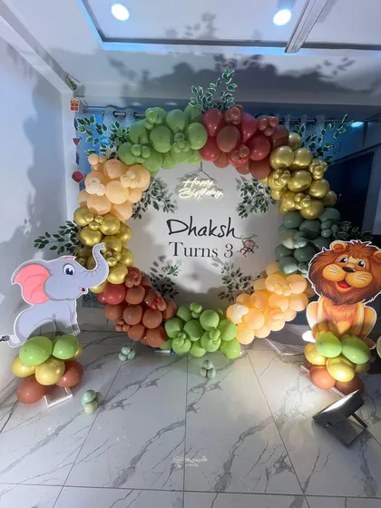 A circular jungle theme backdrop for a third birthday. The balloon wreath is made of various shades of green and orange, with lion and elephant cutouts standing guard.