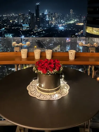 A nighttime view of the romantic table setup. The box of red roses, illuminated by fairy lights, becomes the glowing centerpiece against the sparkling city skyline.
