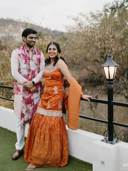 A sweet couple portrait from the mehendi. Their coordinated outfits, with his floral jacket and her orange sharara, look perfect against the outdoor backdrop.