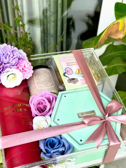 This 'Pastel Delight' acrylic box hamper is a perfect gift for any festivity. It includes an H&M diffuser, a Marks & Spencer photo frame, a scented candle, and delicious treats from Theobroma.