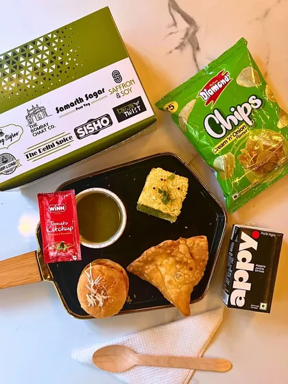 This is a great example of our popular pure veg snack box, starting at just ₹75. It includes a balanced mix of savory items like a samosa and dhokla, a mini bun, chips, a drink, and condiments. It's the perfect light meal for hi-teas and corporate breaks.