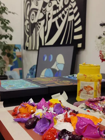 The beautiful chaos of a work-in-progress. A palette of thick, vibrant acrylic paints sits ready on the table, with finished and unfinished canvases in the background, capturing the energy of our creative process.
