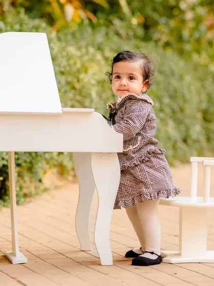 A sweet baby girl in a plaid dress standing at a miniature white piano during her outdoor milestone photoshoot.