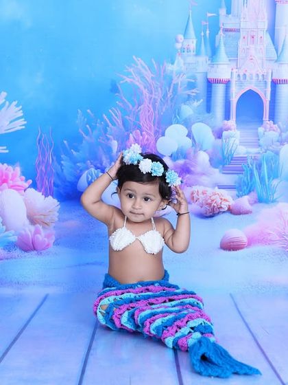 A baby girl in a mermaid costume adjusts her floral crown, looking absolutely adorable. The pastel-colored coral reef and castle create a dreamy and beautiful setting.