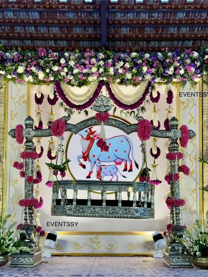 The main stage for a traditional Hindu naming ceremony, featuring an ornate silver-plated cradle. The backdrop is inspired by Pichwai art, depicting Lord Krishna and holy cows, and is framed with floral garlands.