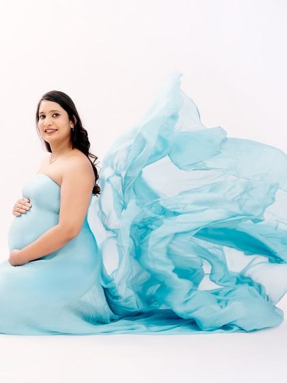 A bright and airy studio portrait. The mom-to-be is kneeling in a light blue gown, with the fabric flowing beautifully behind her against a clean white background.