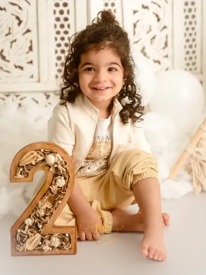 A happy two-year-old boy with curly hair, sitting next to a decorative number "2" for his birthday milestone photos.