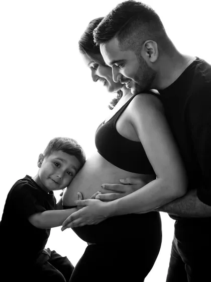 A classic black and white maternity portrait showing the whole family embracing the baby bump. The older brother-to-be gives a gentle hug, creating a timeless and emotional image.