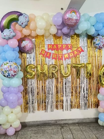 A pastel rainbow theme for a 6th birthday. This at-home decoration features a balloon frame over a tinsel curtain, with rainbow and cloud foil balloons, and the name "SHRUTI" in gold letters.