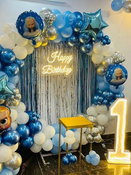 A circular Boss Baby themed backdrop for a first birthday. This home setup includes a blue and silver balloon ring, a shimmer curtain, a neon sign, and a light-up number '1'.