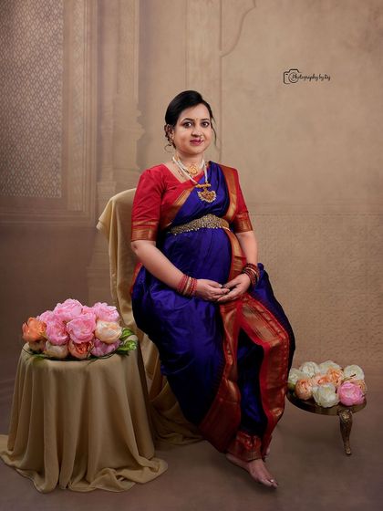 A regal studio portrait of a mother-to-be in a traditional Maharashtrian-style nauvari saree, seated with floral props.