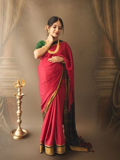 An elegant solo portrait of a mom-to-be in a red saree, standing beside a traditional lamp against a classic painted backdrop.