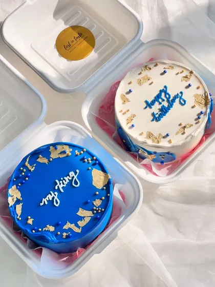 A pair of coordinated bento cakes. One is a solid royal blue with gold leaf, and the other is a white cake with blue script and matching gold accents, creating a beautiful set.