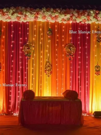 A warm and inviting Mehendi stage with orange and pink drapes, illuminated by fairy lights and decorated with traditional hangings.