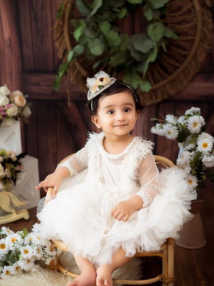 A classic portrait of a baby girl in a white lace dress. The rustic barn-door background and soft lighting create a timeless and elegant look.