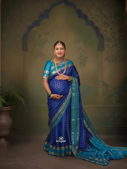 A beautiful portrait of a mother-to-be in a blue checkered silk saree, standing against a classic painted backdrop for a timeless feel.
