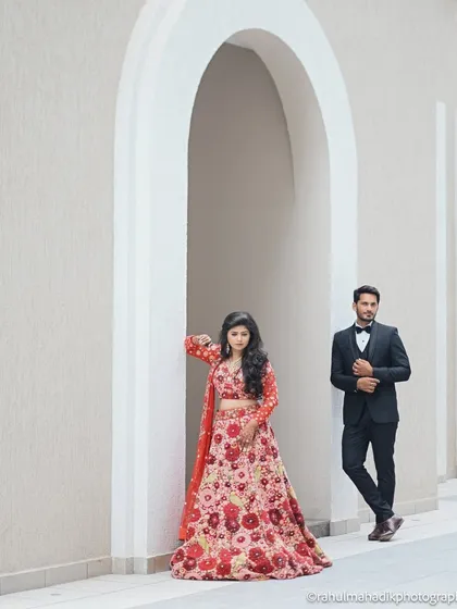 A stylish, minimalist portrait using modern architecture. The couple is framed within a clean white archway, with the bride's vibrant floral lehenga providing a beautiful contrast.