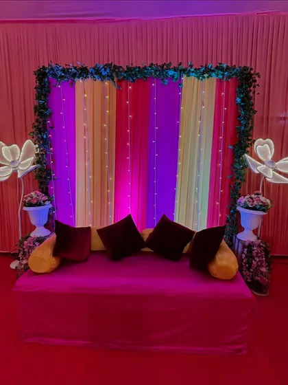The focal point of the Mehendi ceremony is this colorful seating area for the bride. The backdrop features bright striped drapes and fairy lights, framed by a green leaf border and large decorative flowers.