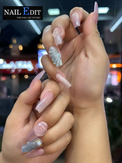 A full view of our long, coffin-shaped nail extensions featuring sculpted seashell art. The design alternates between translucent pink nails and detailed, textured shells with rhinestone and pearl accents for a mermaid-inspired look.