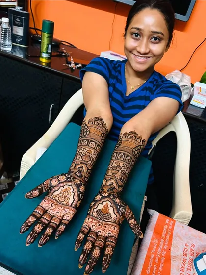 A smiling bride proudly displaying her full bridal mehendi. Both arms are adorned with intricate patterns telling her story, ready for her big day.