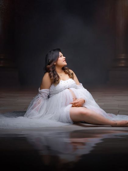 A dreamy and ethereal portrait of a mother-to-be in a white tulle gown. The reflective floor and moody lighting create a magical, water-like effect in the studio.